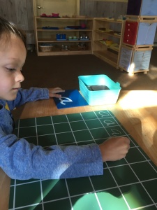 Child practices forming the letter "a" on a gridded chalkboard.
